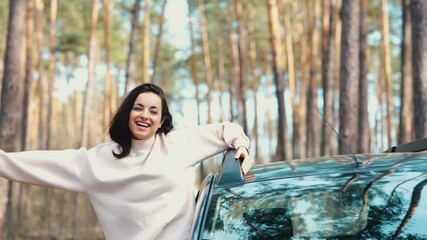 Wall Mural - Young woman sit in car during travelling break. Slow motion of attractive cheerful girl stand out of machine window and scream of happiness. Wave with hands and enjoy riding.