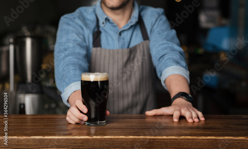 Barman in denim shirt and apron stands behind wooden counter and gives glass of dark beer