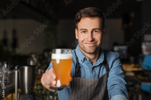 Pint ale. Smiling barman in apron holds out glass of light beer in interior of pub