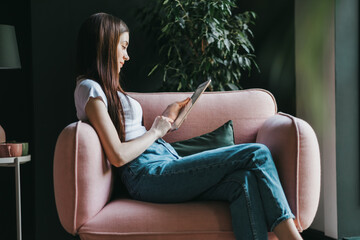 Beautiful young caucasian woman sitting on a pink sofa and using tablet computer. Work at home.