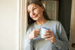 © shurkin_son - Beverage, comfort and daily routine. Attractive gray haired female on retirement staring her day with cup of morning coffee, posing in doorway, holding mug, having dreamy thoughtful facial expression