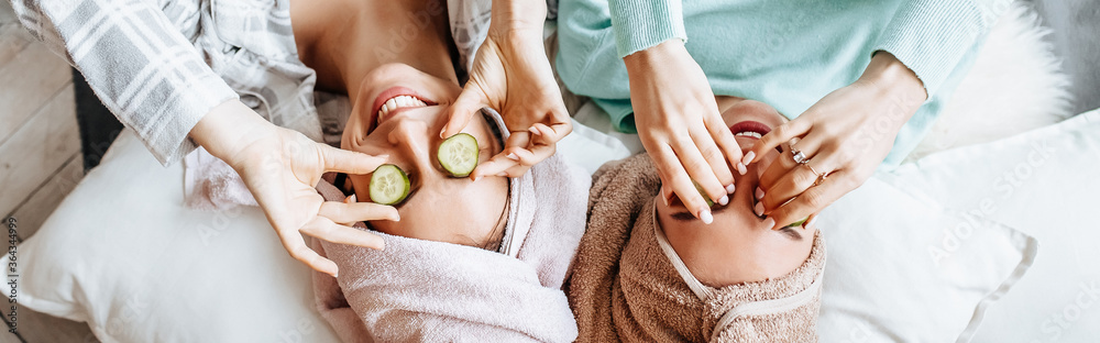 Two girls make homemade face and hair beauty masks. Cucumbers for the freshness of the skin around the eyes. Women take care of youthful skin. Girlfriends laugh at home lying on the floor on pillows. - obrazy, fototapety, plakaty 