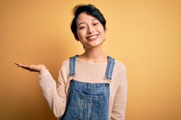 Canvas Print - Young beautiful asian girl wearing casual denim overalls over isolated yellow background smiling cheerful presenting and pointing with palm of hand looking at the camera.