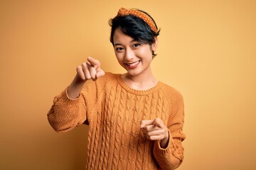 Wall Mural - Young beautiful asian girl wearing casual sweater and diadem standing over yellow background pointing to you and the camera with fingers, smiling positive and cheerful