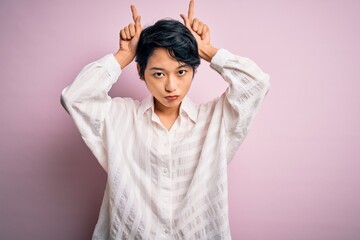 Wall Mural - Young beautiful asian girl wearing casual shirt standing over isolated pink background doing funny gesture with finger over head as bull horns