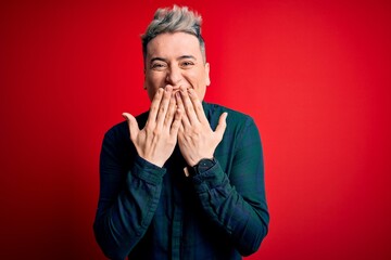 Wall Mural - Young handsome modern man wearing elegant green shirt over red isolated background laughing and embarrassed giggle covering mouth with hands, gossip and scandal concept