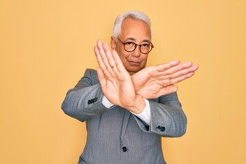 Middle age senior grey-haired handsome business man wearing glasses over yellow background Rejection expression crossing arms and palms doing negative sign, angry face