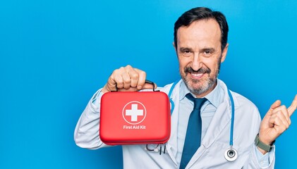 Middle age handsome doctor man wearing coat and stethoscope holding first aid kit smiling happy pointing with hand and finger to the side