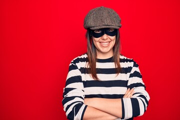 Young beautiful brunette burglar woman wearing cap and mask over isolated red background happy face smiling with crossed arms looking at the camera. Positive person.