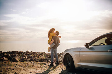 Wall Mural - Romantic couple is standing near a Muscle car on the beach. The handsome bearded man and an attractive young woman have a Love story.