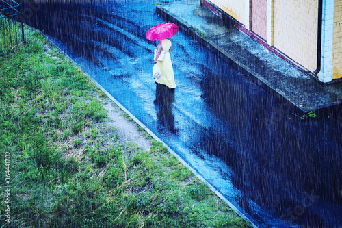 Girl with red umbrella in yellow dress. Wet slippery road. Rain in city. Damp rainy summer weather. Woman with bag. Raindrops are falling. Puddles on the asphalt. Autumn rain.