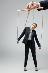 cropped view of puppeteer holding businesswoman marionette on strings isolated on grey