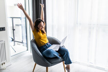 Happy Relaxed Casual Woman Sitting On The Chair With A Laptop In Front Of Her Stretching Her Arms Above Her Head.