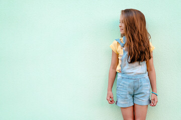 Wall Mural - Portrait of cute blonde girl child in a denim jumpsuit 9-13 years old copy space isolated on pastel background. Adorable female child model posing indoors.