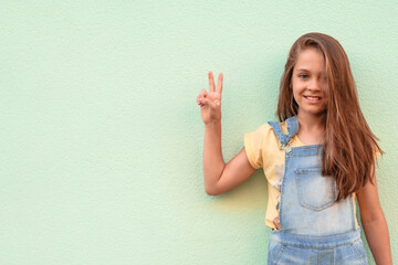 Wall Mural - Portrait of a little girl child in a denim jumpsuit 9-13 years old on an empty wall, the girl shows two fingers symbolizing that everything is good