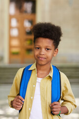 Wall Mural - Vertical waist up portrait of African-American schoolboy carrying backpack and smiling at camera while posing outdoors against school building