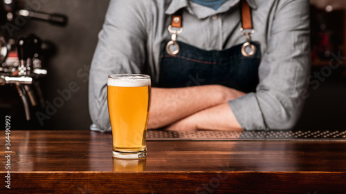Talk with bartender with drink. Man in apron with crossed arms, and light beer with foam on wooden bar counter
