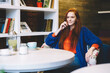 © BullRun - Smiling young woman with read-head sitting in comfortable stylish cafeteria enjoying recreation indoors.Cheerful foxy teenager dressed in casual outfit spending free time in coffee shop interior