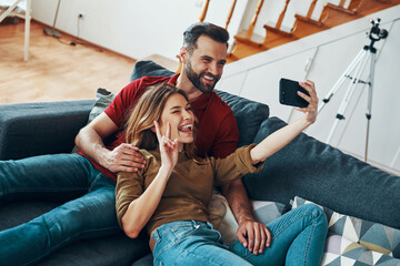 Canvas Print - Playful young couple in casual clothing bonding together and smiling while taking selfie indoors