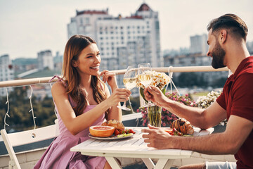 Sticker - Charming young couple in casual clothing toasting each other and smiling while sitting on the rooftop patio outdoors