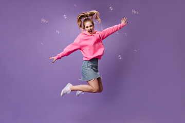 Cool girl in denim skirt and hoodie jumping on purple background. Smiling teen in pink sweatshirt has fun on isolated backdrop