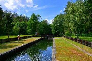 View of a narrow channel with two concrete edges on both sides, a small, neatly mown lawn and some wooden dam door in the background located next to a dense forest or moor seen on a cloudy summer day