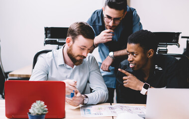 Multicultural male colleagues dressed in formal wear discussing productive strategy of developing common startup project collaborating with each other at modern laptop device in stylish office