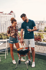 Sticker - Full length of young couple in casual clothing preparing barbecue and smiling while standing on the rooftop patio outdoors