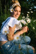 Side View Of Young Woman Looking At Camera In Casual Clothes Enjoying Smell Of Fresh Chamomiles While Collecting Flowers On Green Meadow In Spring