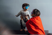 Side View Of Playful Woman In Superhero Mask And Cloak Tossing Little Boy While Spending Time Together At Home And Having Fun