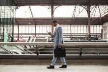 Side View Of Stylish African-American Businessman With Suitcase Walking On Station And Watching Smartphone.