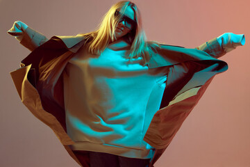 Stylish young girl, cool dancing in a hoodie, sunglasses and developing cloak, on a neon background.