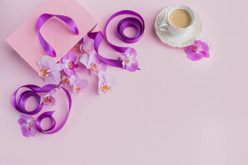 Wall Mural - Flower overhead composition on light pink background top view. Cup of coffee, pink gift bag with purple ribbons and pink orchid flowers.