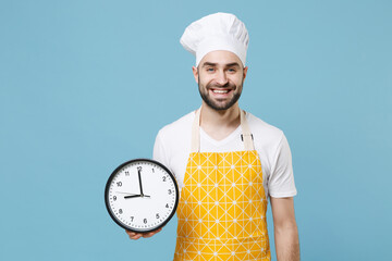 Wall Mural - Smiling young bearded male chef or cook baker man in apron white t-shirt toque chefs hat isolated on pastel blue wall background studio portrait. Cooking food concept. Mock up copy space. Hold clock.