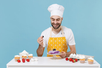 Funny male chef or cook baker man in apron white t-shirt toque chefs hat cooking at table isolated on blue background. Cooking food concept. Mock up copy space. Decorating dessert cake with berries.