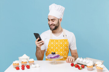 Smiling young bearded male chef or cook baker man in apron white t-shirt toque chefs hat cooking at table isolated on blue background. Cooking food concept. Mock up copy space. Using mobile phone.