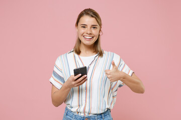 Wall Mural - Smiling young blonde woman girl in casual striped shirt posing isolated on pastel pink background studio portrait. People lifestyle concept. Mock up copy space. Using mobile phone, showing thumb up.