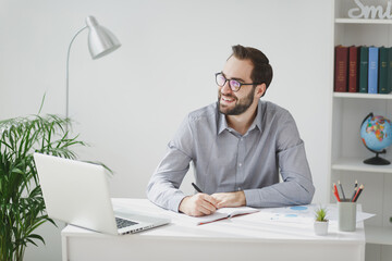 Smiling young bearded business man in gray shirt glasses sit at desk work on laptop pc computer in light office on white wall background. Achievement business career concept Writing notes in notebook.