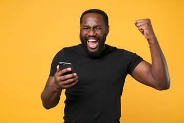 Canvas Print - Happy african american man football fan in casual black t-shirt isolated on yellow background studio portrait. People lifestyle concept. Mock up copy space. Using mobile phone doing winner gesture.