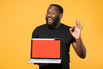 Wall Mural - Excited african american man in casual black t-shirt isolated on yellow background. People lifestyle concept. Mock up copy space. Hold laptop pc computer with blank empty screen showing OK gesture.