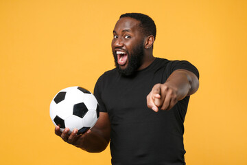 Wall Mural - Angry african american man football fan in black t-shirt isolated on yellow background. Sport family leisure concept. Cheer up support favorite team with soccer ball pointing index finger on camera.