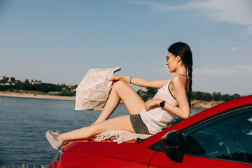 Wall Mural - Young woman lying on car hood, holding a map, looking towards a lake. Concept of tourism.