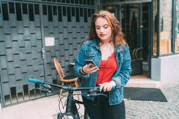 Wall Mural - Portrait of red curled long hair caucasian teen girl on the city street walking with bicycle using the smartphone with earphones. Natural people beauty urban life concept image.