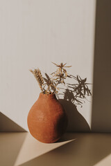 Wall Mural - Red handmade clay flower pot with dry wheat / rye bouquet in sunlight shadows on white background. Minimal modern interior decoration concept.