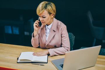 Wall Mural - Mature businesswoman checking notes in notepad while calling on smartphone working at modern laptop computer.Experienced female professor talking on telephone sitting at notebook and netbook