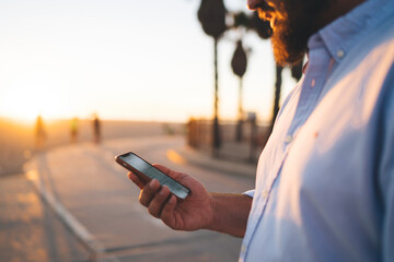 Fototapete - Cropped image of bearded male holding mobile phone reading news information from browsed web page standing on street,hipster guy viewing photos in social networks blogging online via smartphone