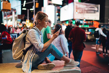 Young Woman Reading Message On Mobile Phone Using Internet In Roaming During Trip To New York, Female Traveler Posting Pictures In Blog Via Smartphone Sitting At Crowded Times Square In Evening