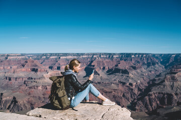 Wall Mural - smiling girl with rucksack using mobile connection for sending messages from rocky cliff in national