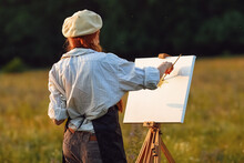  A Girl Artist With Long Red Hair Draws On An Easel With A Brush Against The Background Of The Sunset