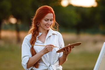  a girl artist with long red hair draws on an easel with a brush against the background of the sunset